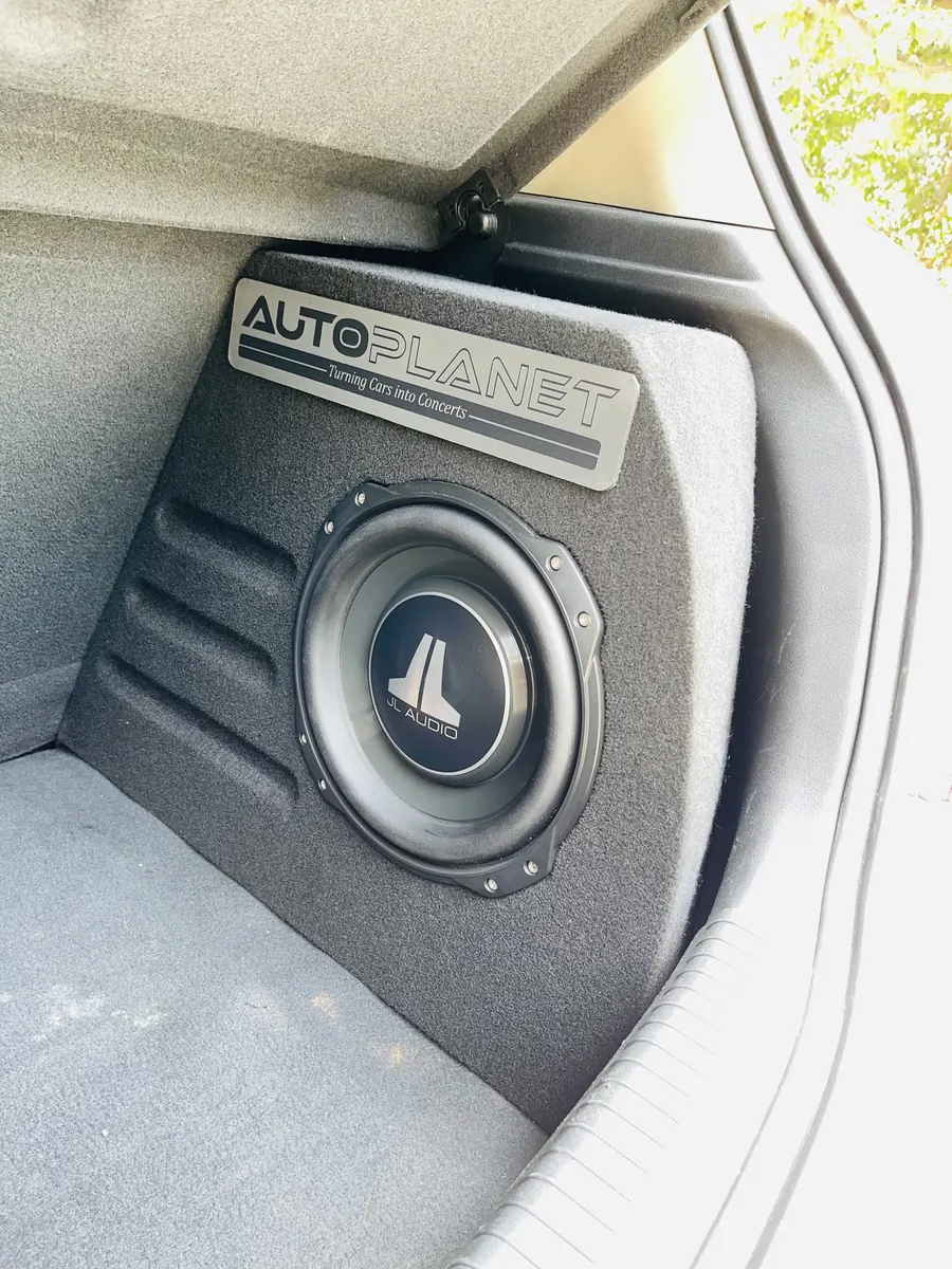 A JL Audio subwoofer flush-mounted into a fabricated carpet-trimmed enclosure carrying an etched Auto Planet plate