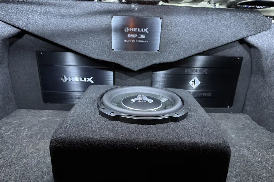 A black-carpeted boot build: a Helix DSP.3S badge plate flanked by two Helix amplifiers behind a fabricated panel, with a JL Audio subwoofer mounted in a carpeted enclosure in the foreground.