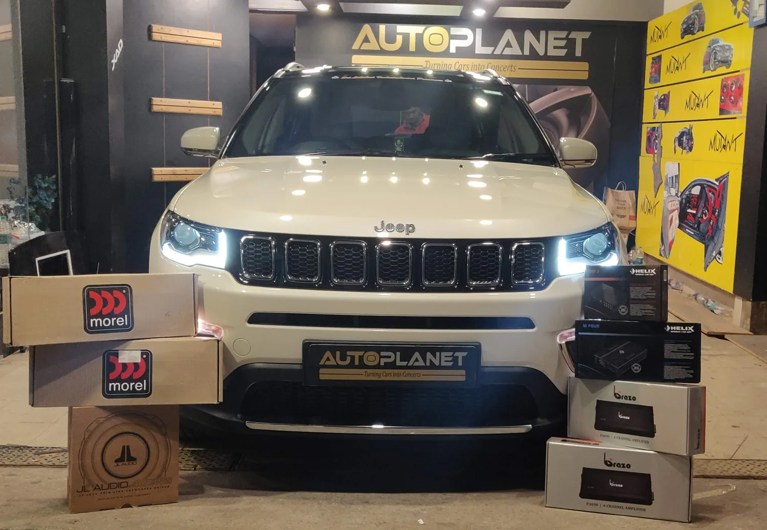 A white Jeep Compass in the Auto Planet showroom, headlights on, with Morel, JL Audio, Helix and Brazo boxes stacked in front of it