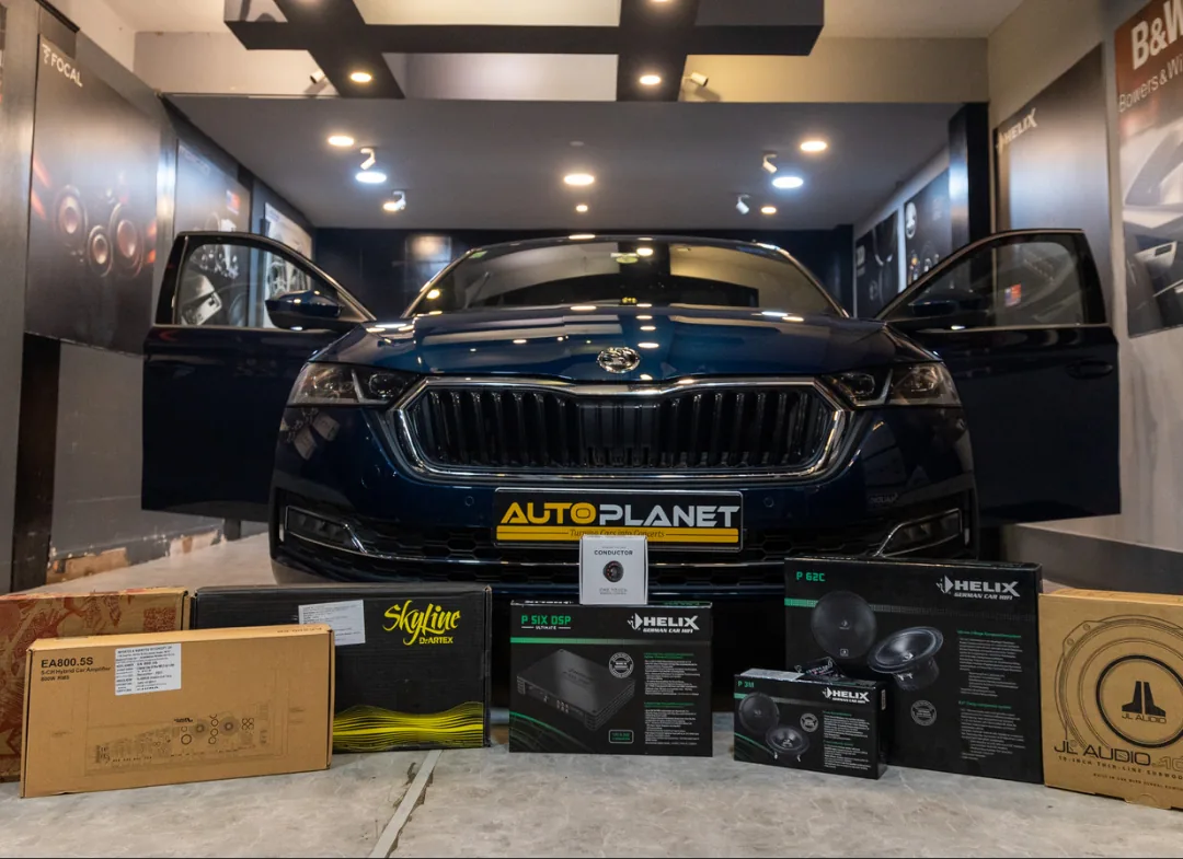 A dark blue Skoda in the Auto Planet showroom with its doors open, Helix, JL Audio and D-Artex boxes lined up on the floor in front of it