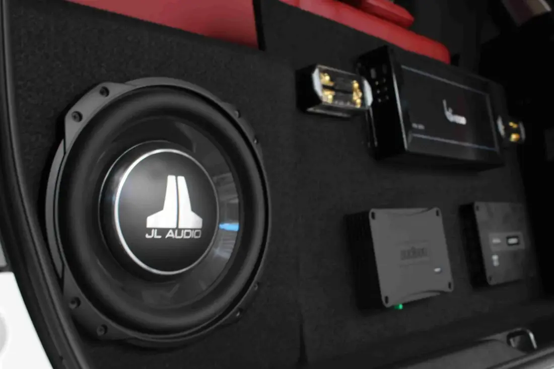 A JL Audio subwoofer and matched amplifiers set into a black-carpet boot panel, red interior trim behind